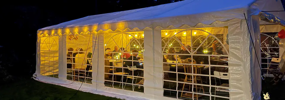 Partytelt i en hage om kvelden, der lysslynger lyser opp innsiden og gjestene ved bordene
