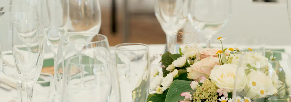 Dekket bord i et partytelt med vinglass, champagneglass, vannglass og blomster på hvit duk