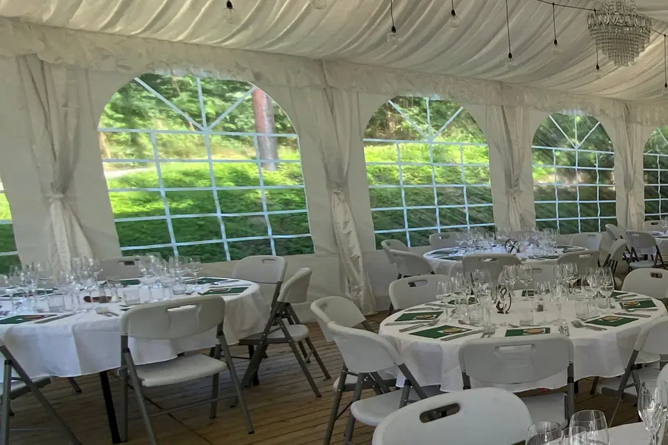 Hvite draperinger knyttet rundt teltbeina mellom vinduene i et partytelt, med lining i taket og runde bord
