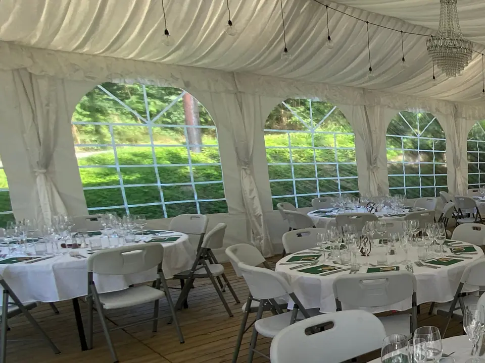 Hvite draperinger knyttet rundt teltbeina mellom vinduene i et partytelt, med lining i taket, runde bord og tregulv