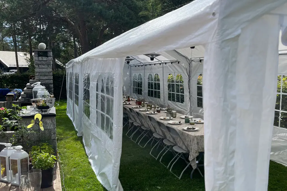Langt, smalt partytelt på plenen med åpen side og et dekket langbord med hvite klappstoler inne