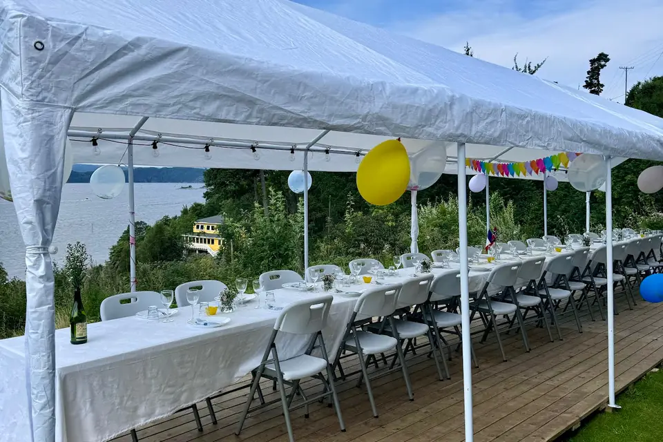 Hvitt partytelt over et langt dekket bord på tregulv, pyntet med ballonger og vimpler og med utsikt mot sjøen