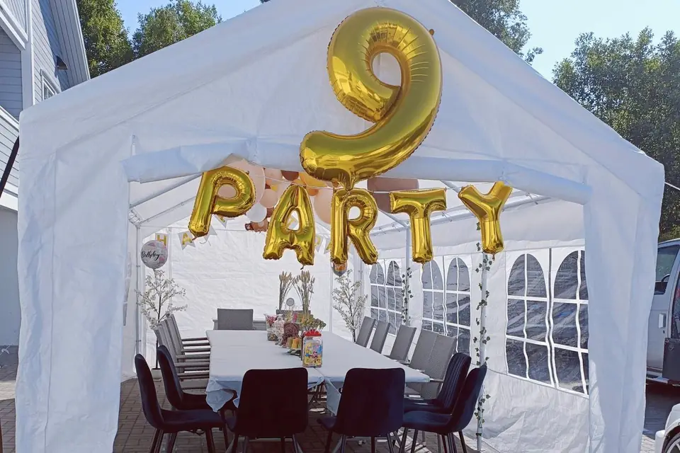 Hvitt partytelt på en gårdsplass, pyntet med gullballonger til bursdag og med dekket langbord og stoler inne