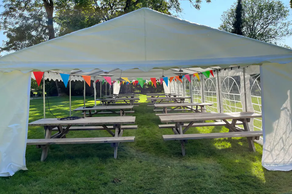 Partytelt med åpen gavl og rader av piknikbord under fargerike vimpler på plenen ved Carlberg Gård