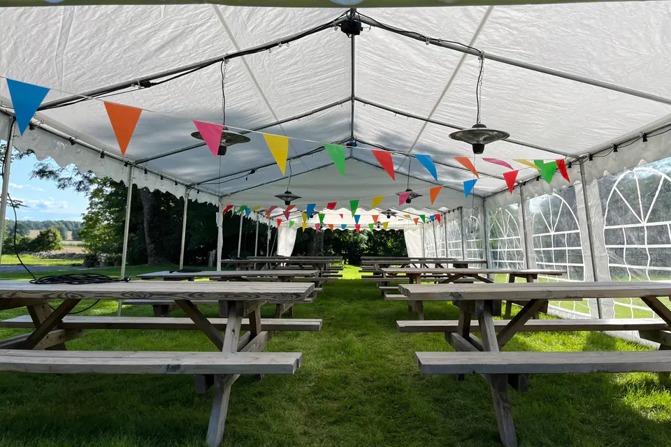 Innsiden av et langt partytelt med piknikbord, varmelamper og fargerike vimpler under taket