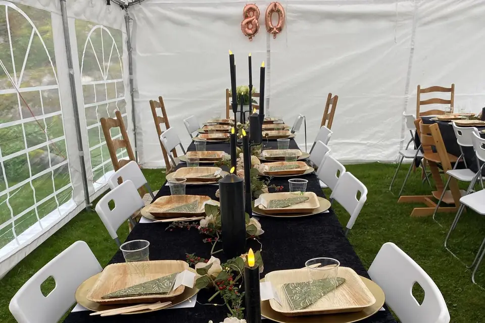 Langbord med sort duk, tallerkener av palmeblad og hvite klappstoler i et partytelt pyntet til 80-årsdag