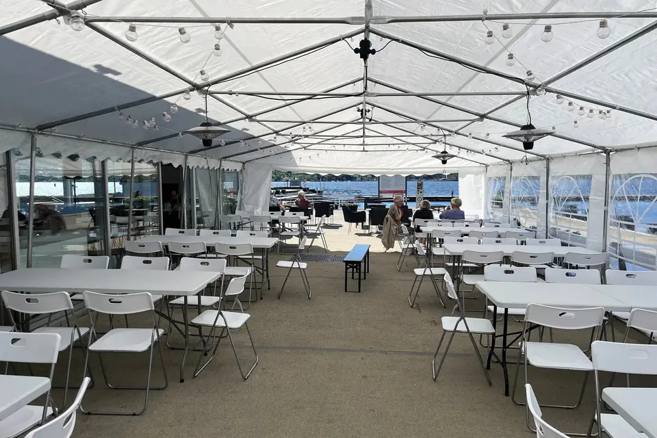Innsiden av et langt partytelt ved Cafe Sjøstjernen med hvite bord og klappstoler, lyslenker og terrassevarmere i taket, og sjøen synlig gjennom den åpne gavlen