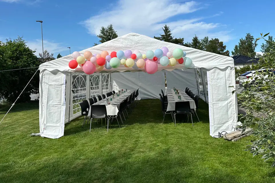 Hvitt partytelt på plenen med ballongbue i pastellfarger over inngangen og to dekkede langbord inne i teltet