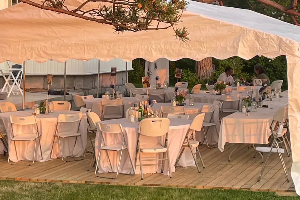 Partytelt med åpne sider i gyllent kveldslys, med runde bord med hvite duker, klappstoler, blomster og lys på et tregulv under en furu