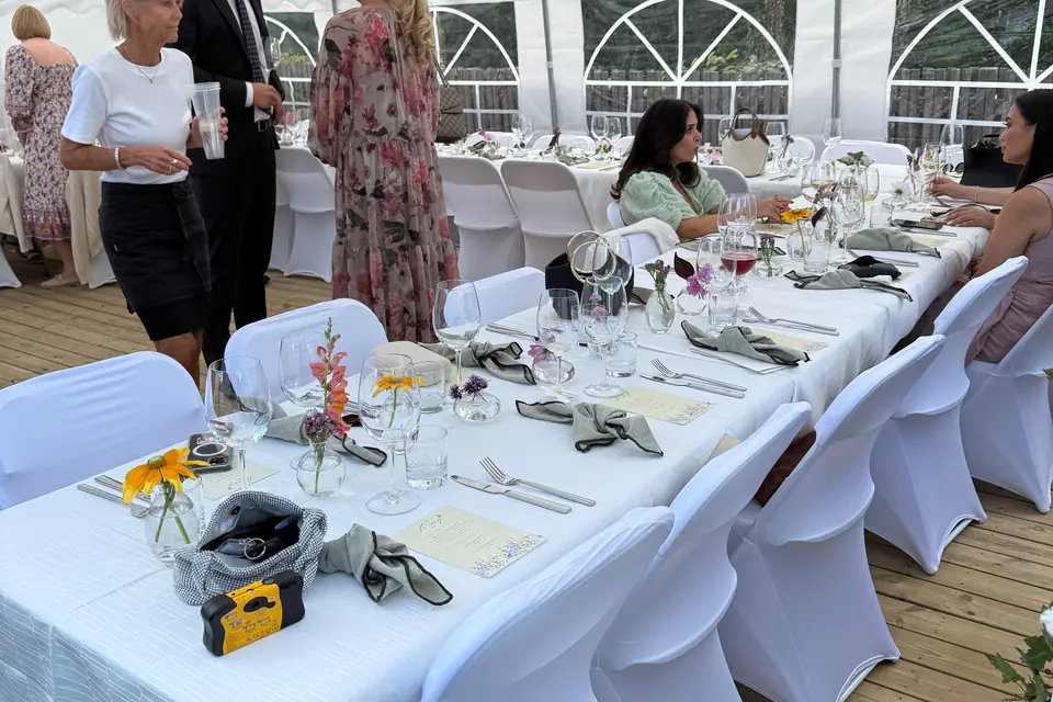 Bryllupsbord i partytelt med hvite duker, hvite stoltrekk, vinglass og små blomstervaser, med teltvinduer og gjester i bakgrunnen