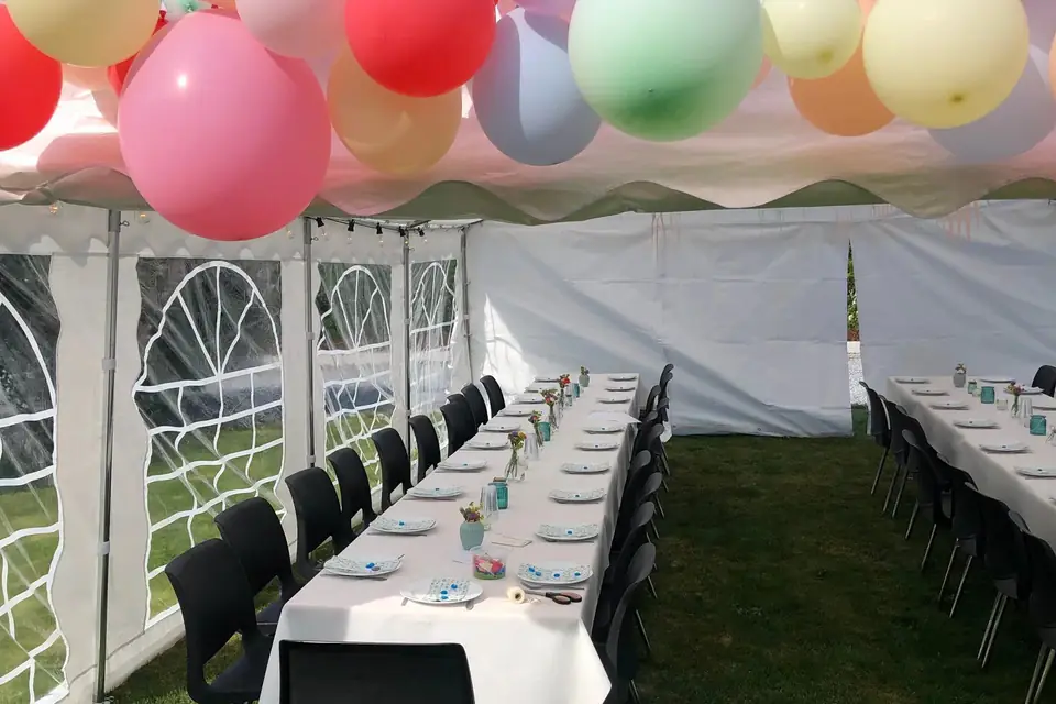 Ballonger i pastellfarger langs teltkanten over to dekkede langbord med svarte stoler inne i et hvitt partytelt
