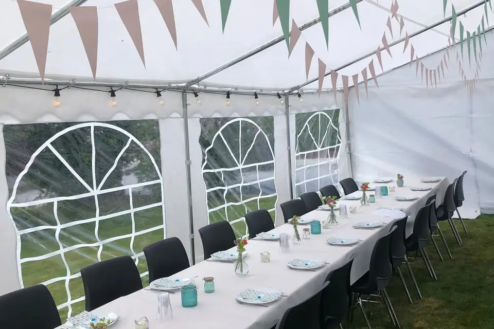 Langbord med hvite duker, sorte stoler og små blomstervaser inne i et partytelt med vindusvegger, lyslenke og vimpler i taket