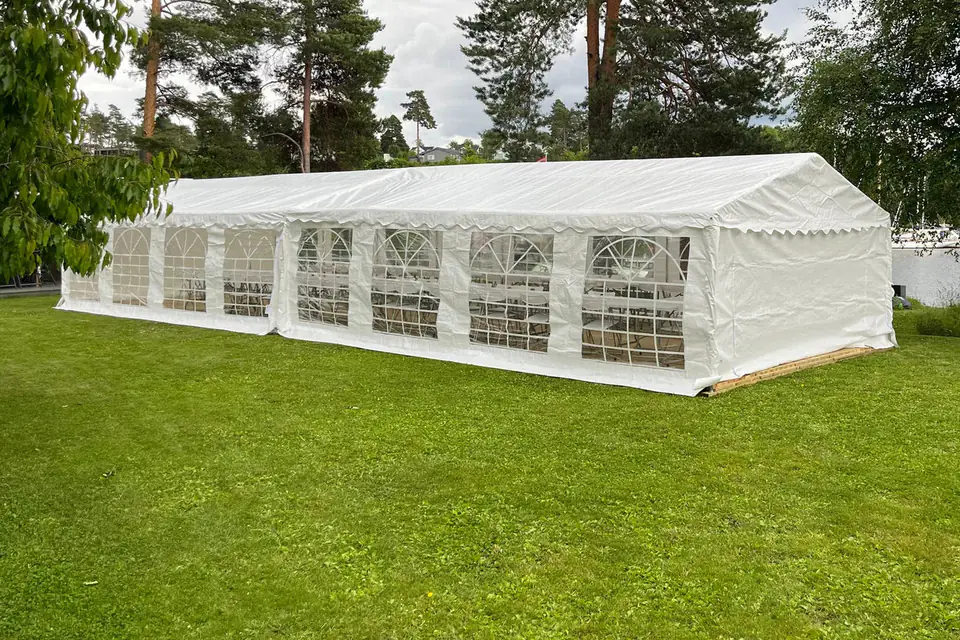 Langt hvitt partytelt med buede vinduer i sideveggene, satt opp på en gressplen ved vannet mellom høye furutrær