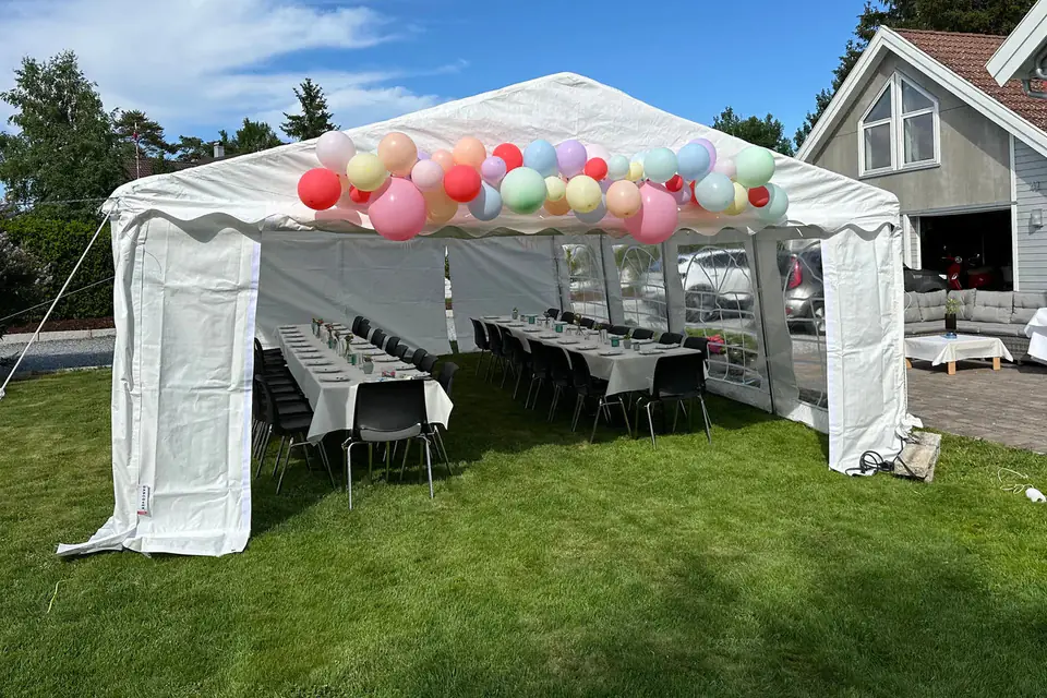 Partytelt med åpen gavl og ballongbue over inngangen, dekket med to langbord på plenen ved et bolighus en solrik dag