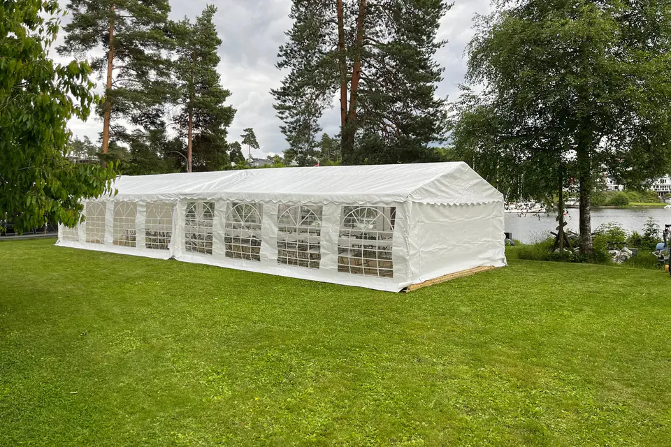 Partytelt med vindusvegger på en plen ved vannet, med høye furutrær rundt
