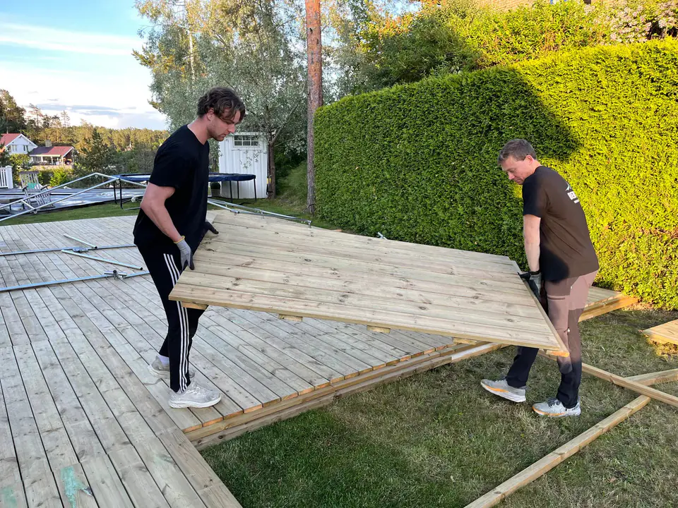 To montører fra Berg Event bærer en gulvmodul av terrassebord på plass i en hage