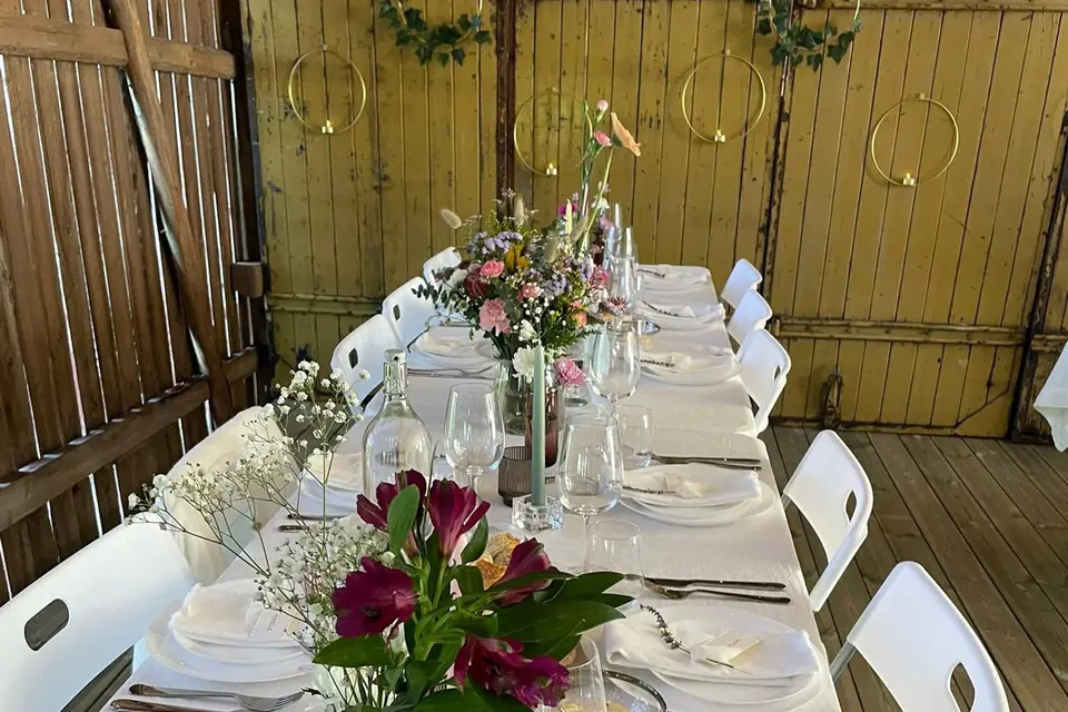 Hvite klappstoler langs et dekket langbord med blomster i en låve