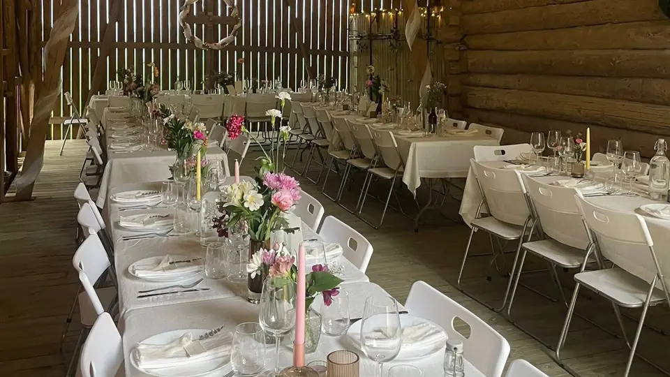 Lange, dekkede bord med hvite klappstoler i en låve pyntet med blomster og lys