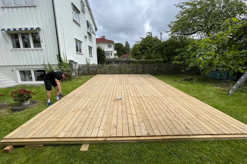 Ferdig tregulv av terrassebord på en plen ved et hvitt hus, med en montør i bakgrunnen