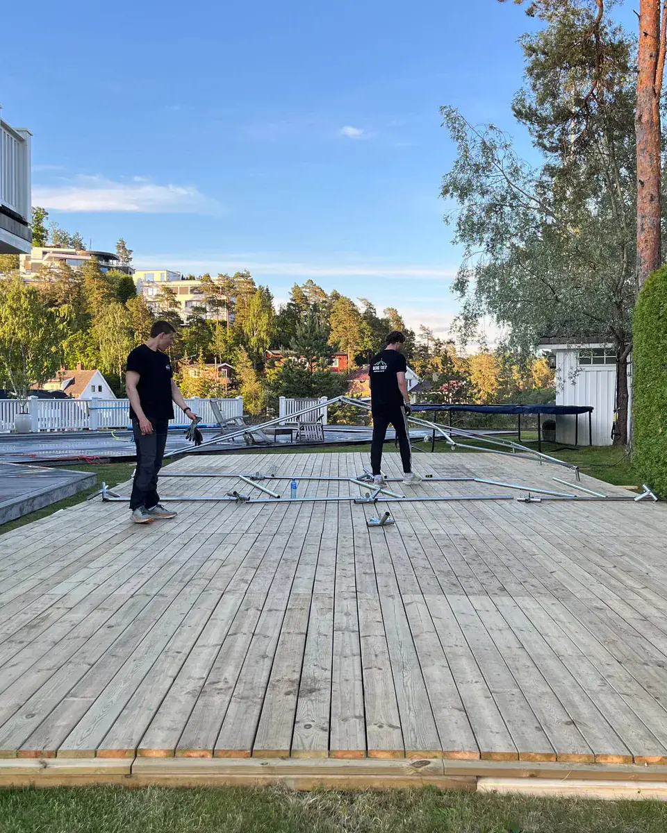 To montører legger ut teltrammen på et stort tregulv i en hage